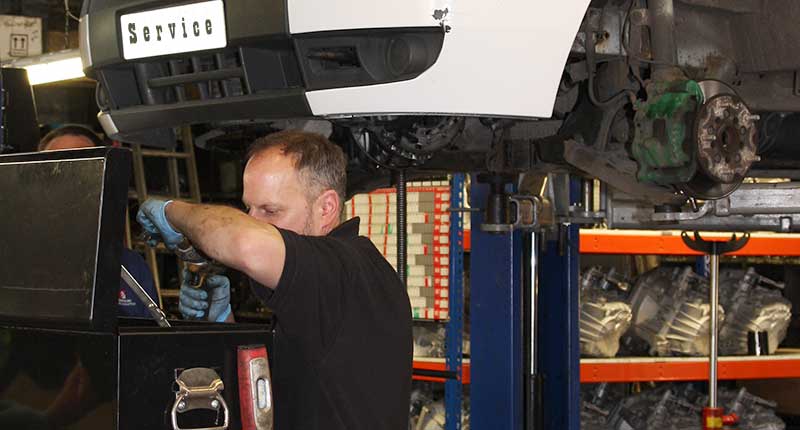 front right corner photo of a mt gear serviceing mechanic at his toolbox with a white volkswagon golf behind him in the air on a ramp at shoulder height with no front wheel on and the brakes being replaced as part of the vehicles service plan