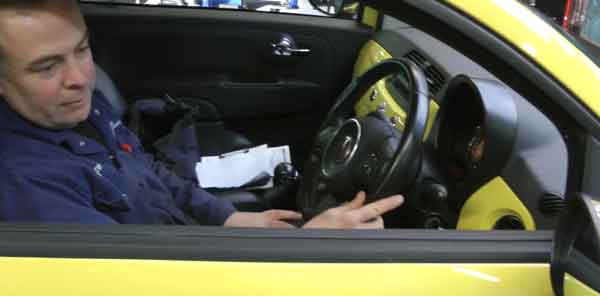 close up side view of an mt gear mechanic driving in a reverse a yellow fiat into the sheffield garage for repairs with one hand on the steering wheel and the other on the gear stick and looking in the cars drivers side door mirror