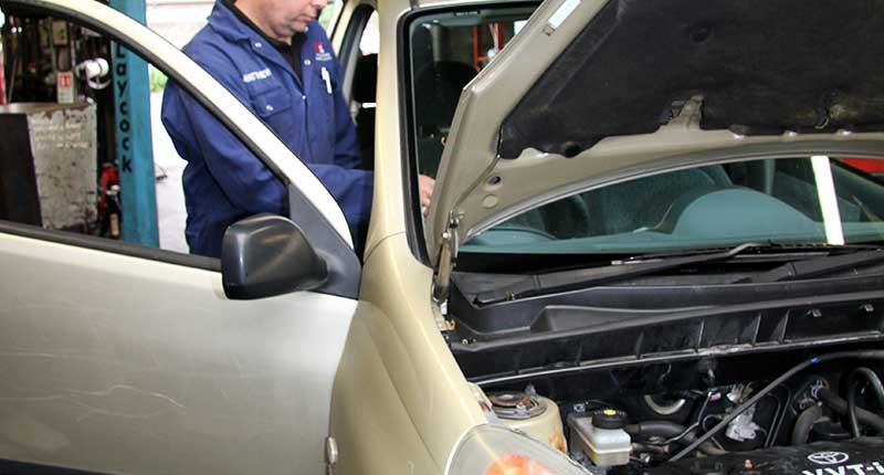 front left corner pgoto of mt gear mot tester testing a cream coloured fiat 500 with one hand on the steering wheel and the other checking the cars lights are working correctly